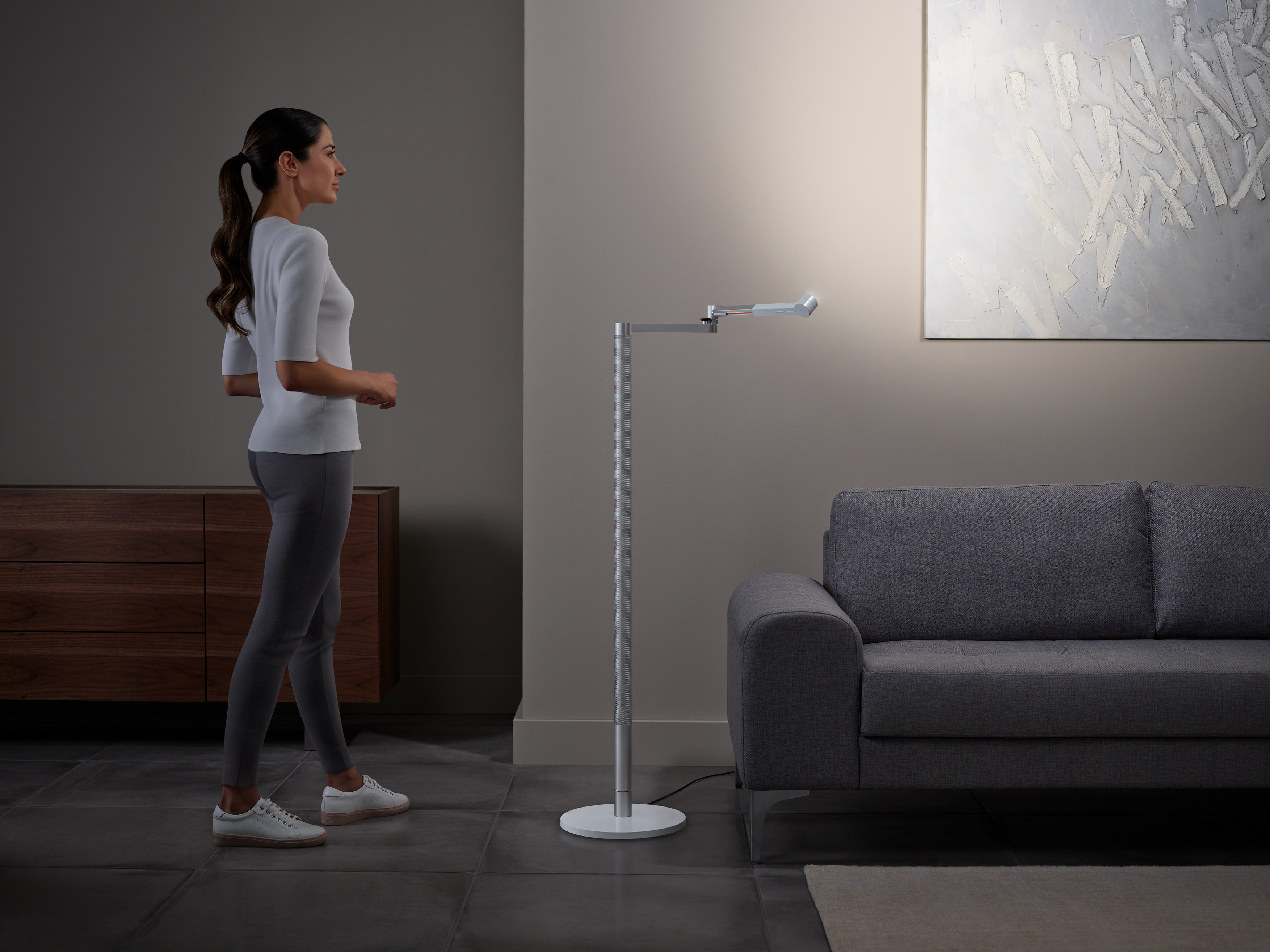 Eine Frau steht in einem sanft beleuchteten Wohnzimmer in der Nähe einer modernen Stehlampe und eines grauen Sofas.