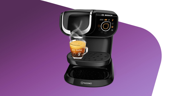 Eine schwarze Kaffeemaschine, die Kaffee in eine Glastasse brüht. Dampf steigt auf.