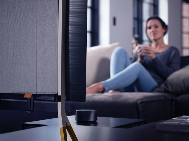 Eine Frau auf einer Couch hält eine Tasse, in der Nähe eines Fernsehers. Ein kleines schwarzes Gerät steht auf dem Tisch. Der Fernseher hat einen weißen und goldenen Ständer.