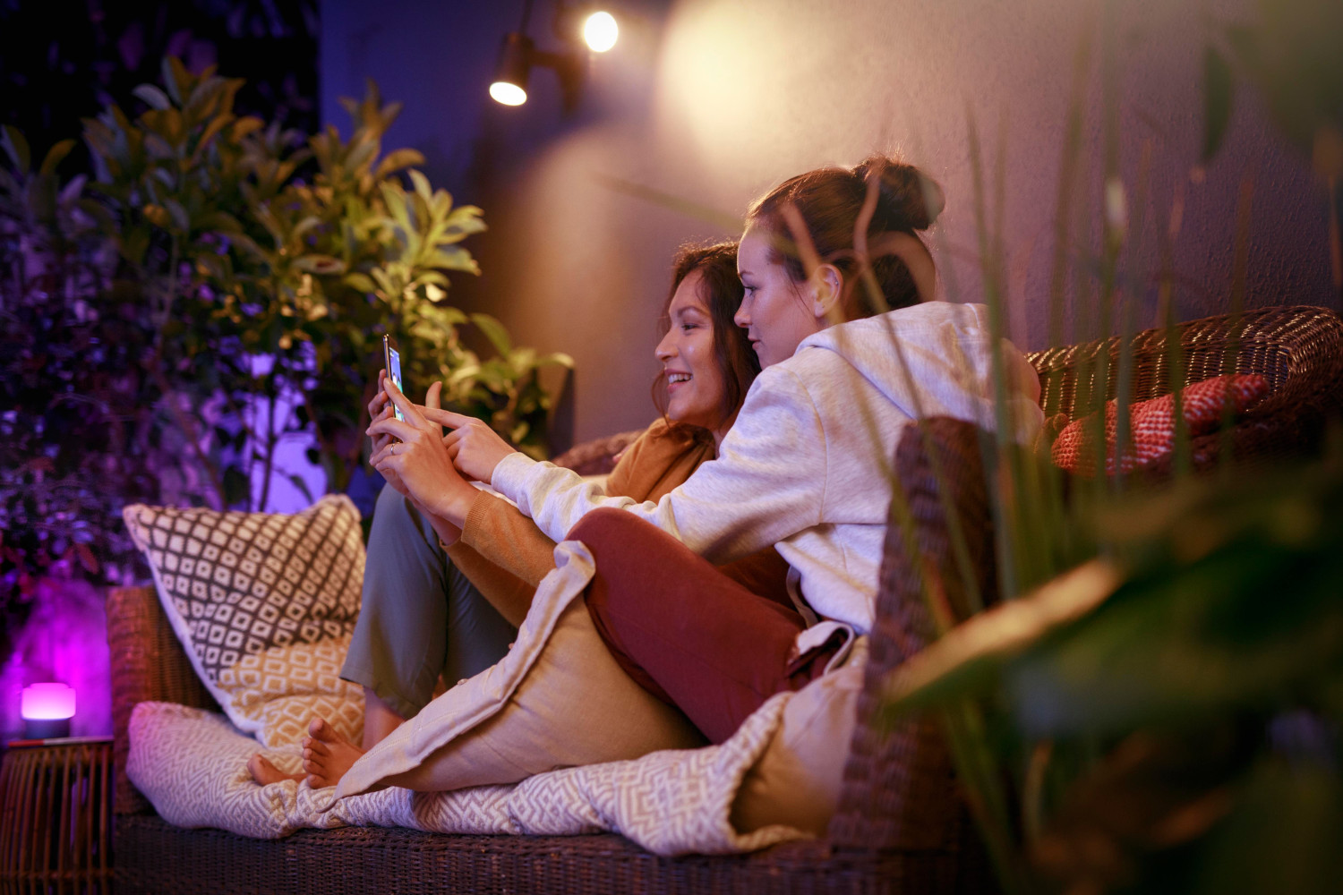 Two women looking at a phone together on a couch in a backyard at night, with lights.