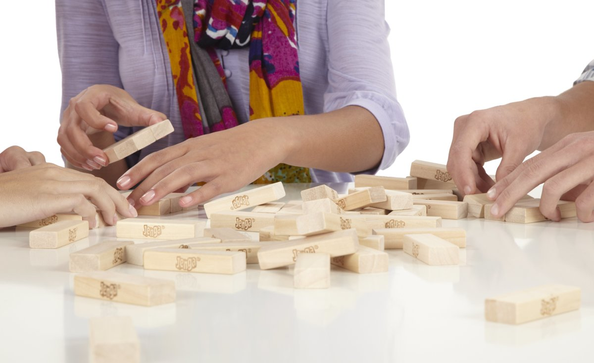 Three people playing Jenga. Wooden blocks are spread on a white table, hands touching blocks.