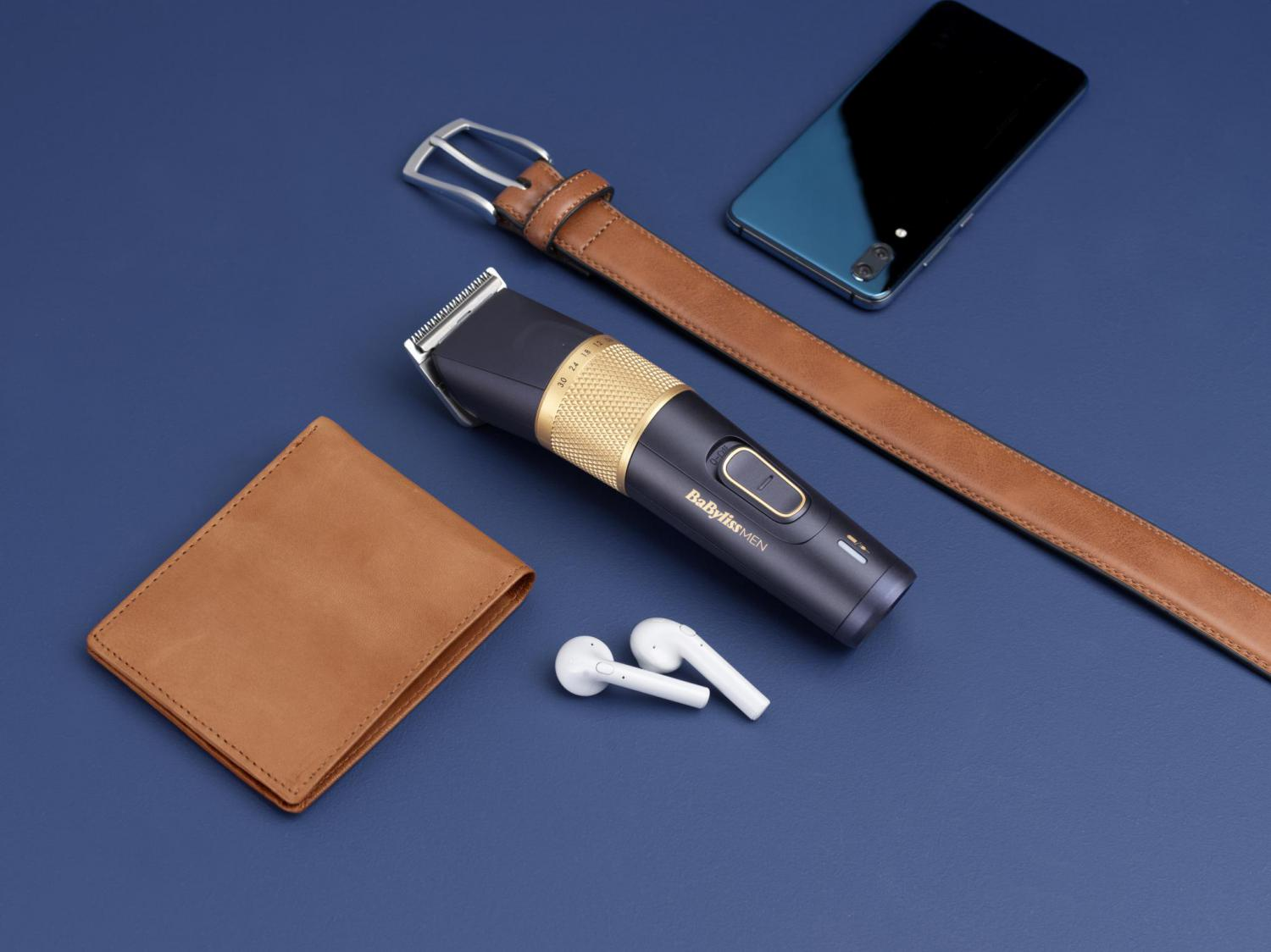 Disposizione di articoli da uomo: trimmer, portafoglio, cintura, telefono e auricolari su superficie blu.
