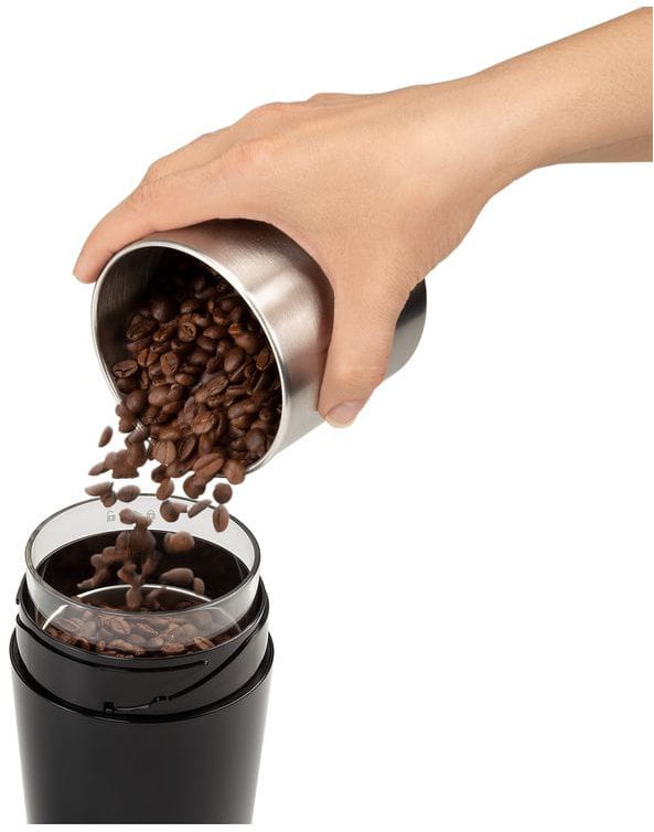 A hand pours coffee beans from a steel container into a black coffee grinder.