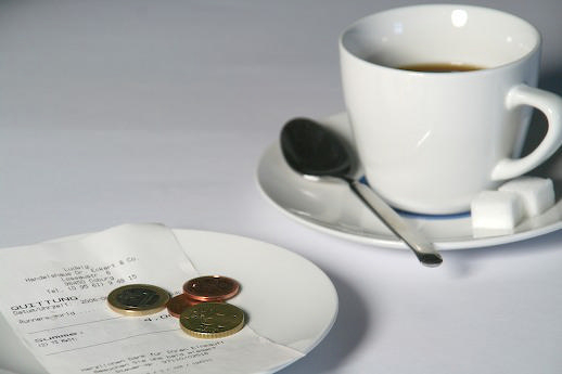 Eine weiße Tasse und Untertasse mit Löffel und Würfelzucker, sowie eine Quittung und Kleingeld auf einem weißen Teller.