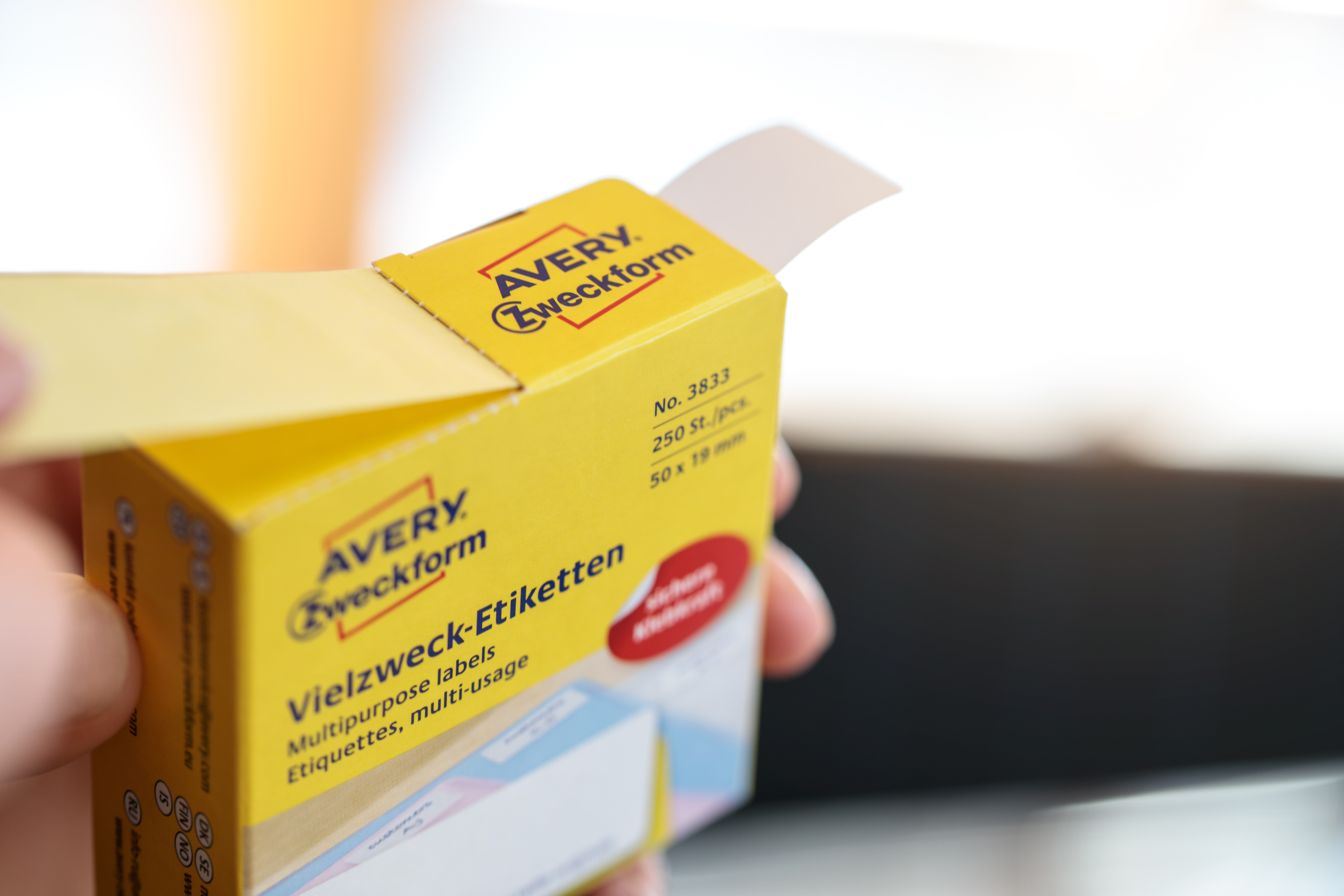 Eine Hand hält eine gelbe Avery Zweckform Etikettenbox. Die Box ist geöffnet und zeigt Etiketten. Der Hintergrund ist unscharf.