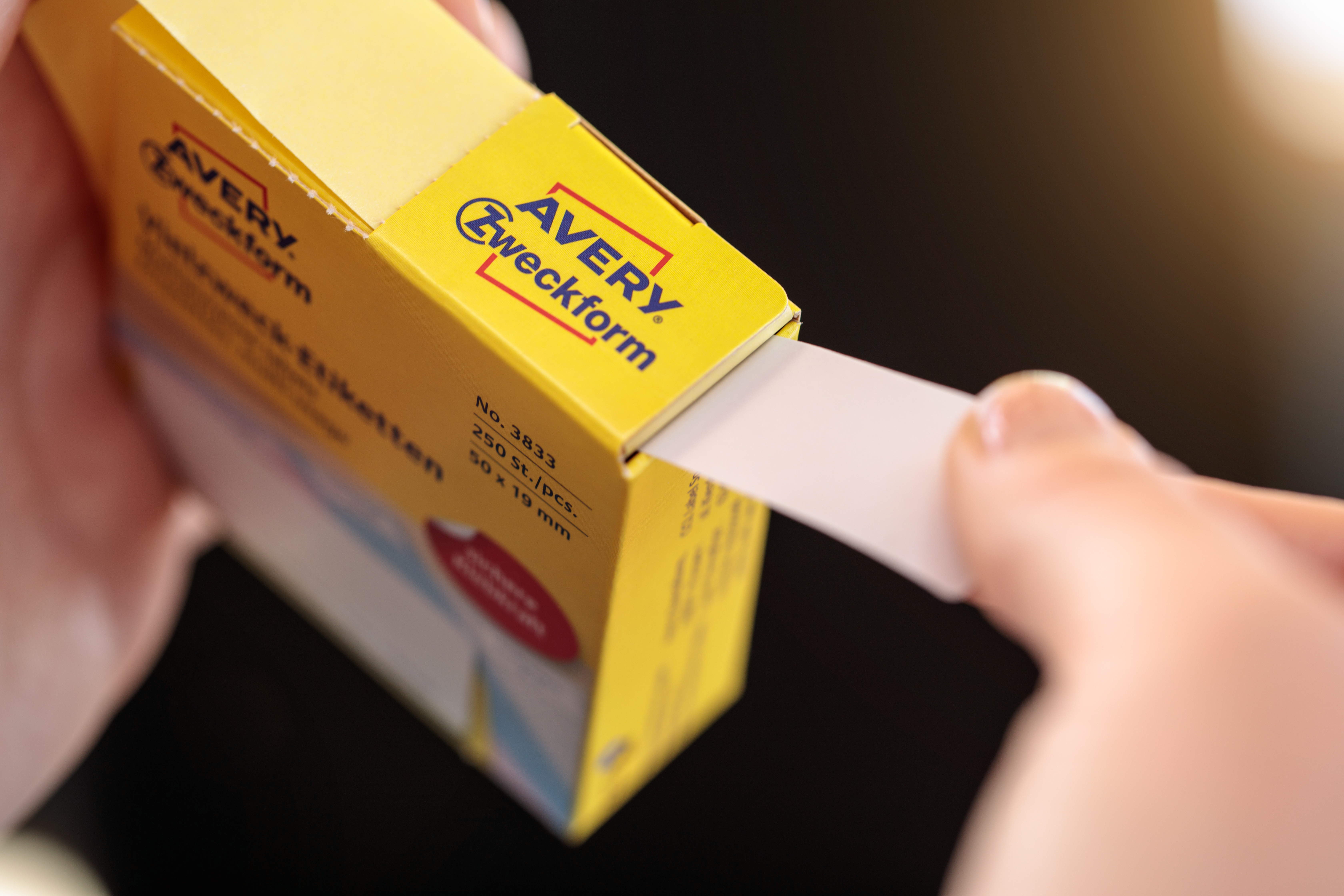 Eine Hand zieht ein weißes Etikett aus einer gelben Schachtel mit der Aufschrift 'AVERY Zweckform'. Der Hintergrund ist unscharf.
