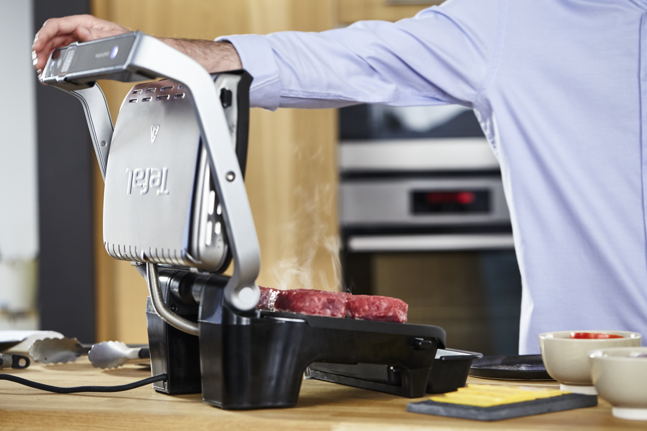 A man grilling burgers with a Tefal grill. Steam rises from the patties. Kitchen setting with wooden counter.