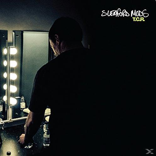 Mann im Schatten steht an einem Spiegel mit Lichtern. Sleaford Mods, T.C.R. ist geschrieben.