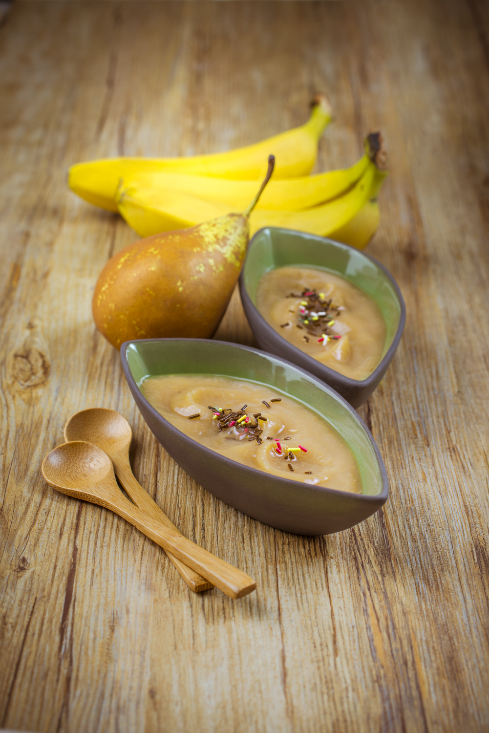 Fruit, banana, pear, and two bowls of blended food with sprinkles, on a wooden table.