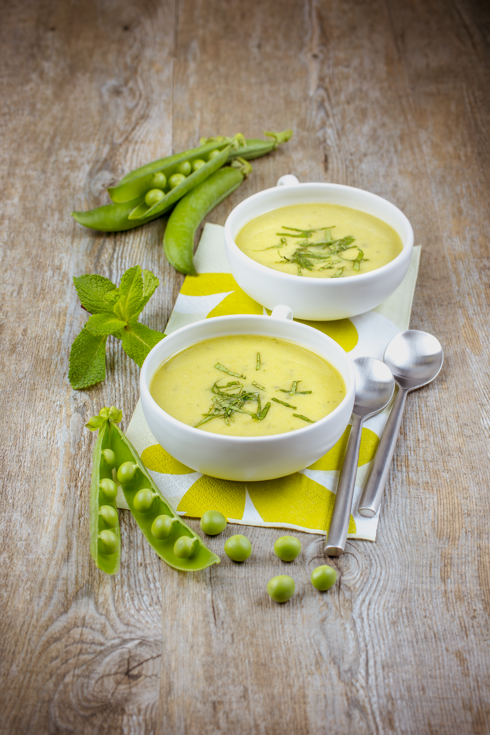Two bowls of soup with spoons and pea pods on a wooden table.