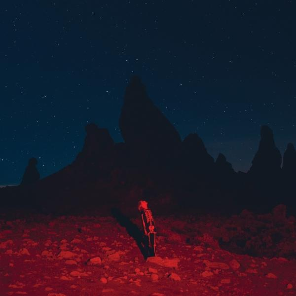 Eine karge Landschaft bei Nacht mit einer Skelettfigur, die von rotem Licht beleuchtet wird, Sterne am Himmel.