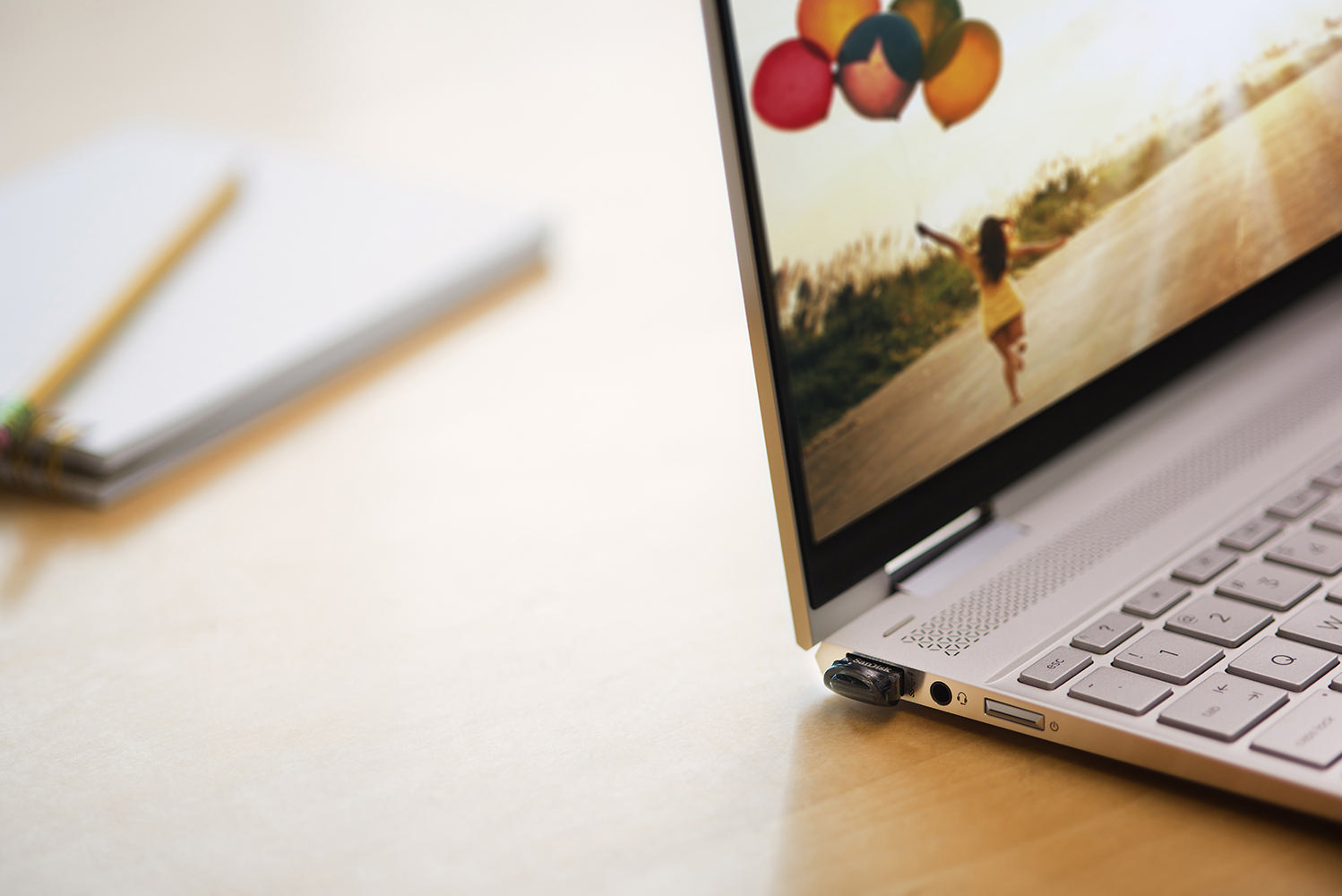 Ein silberner Laptop mit einem Bildschirm, der ein Foto eines Mädchens mit Ballons zeigt, neben Notizblock und Bleistift.