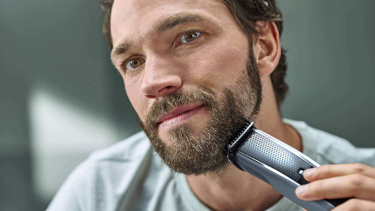 Een man scheert zijn baard met een trimmer. Hij heeft bruin haar en een volle baard. De trimmer is zwart en grijs.