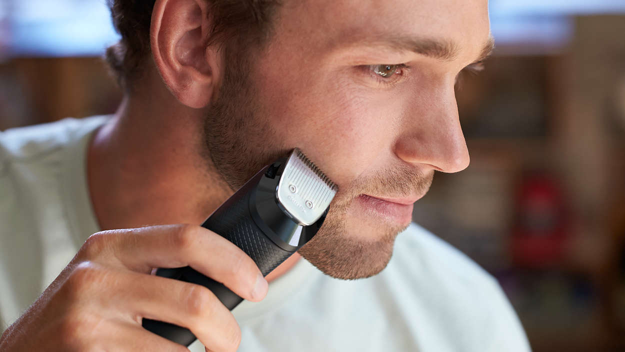 Een man scheert zijn baard met een elektrische trimmer. Hij heeft een lichte huid en de trimmer is zwart met een zilveren mes.
