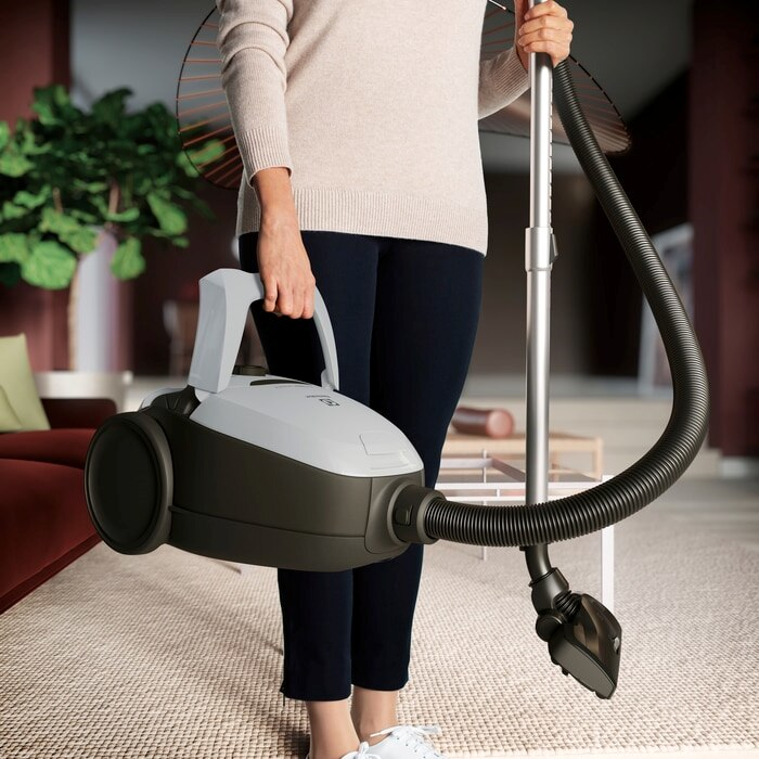 Nő porszívót tart. A porszívó szürke és fekete, tömlővel és kefével.