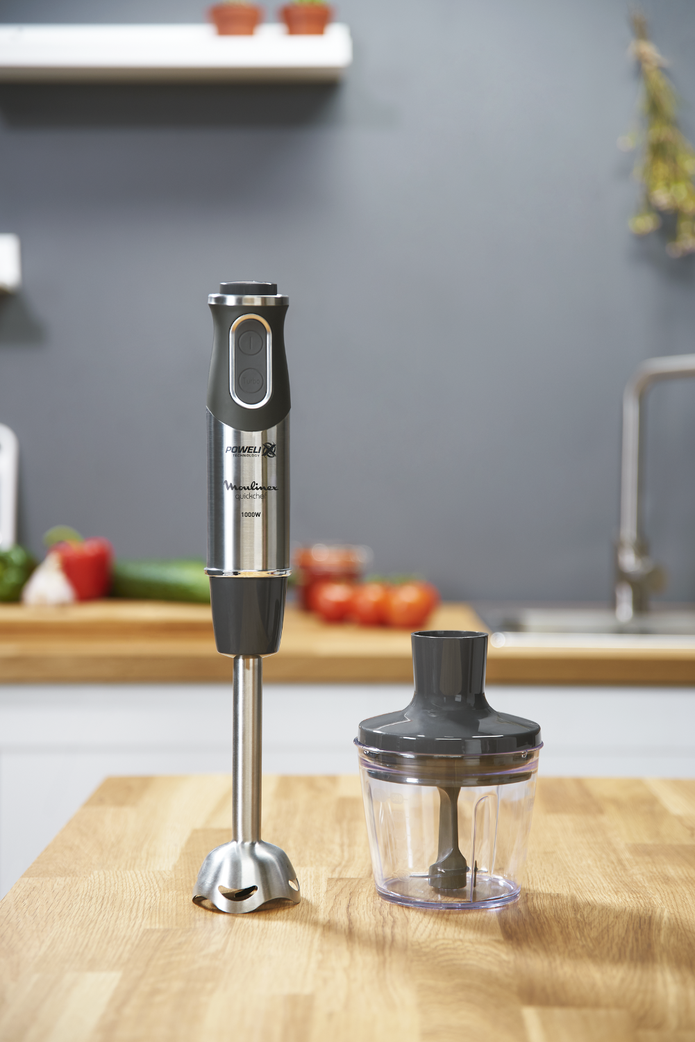 A silver and black Moulinex hand blender with a clear container on a wooden countertop.