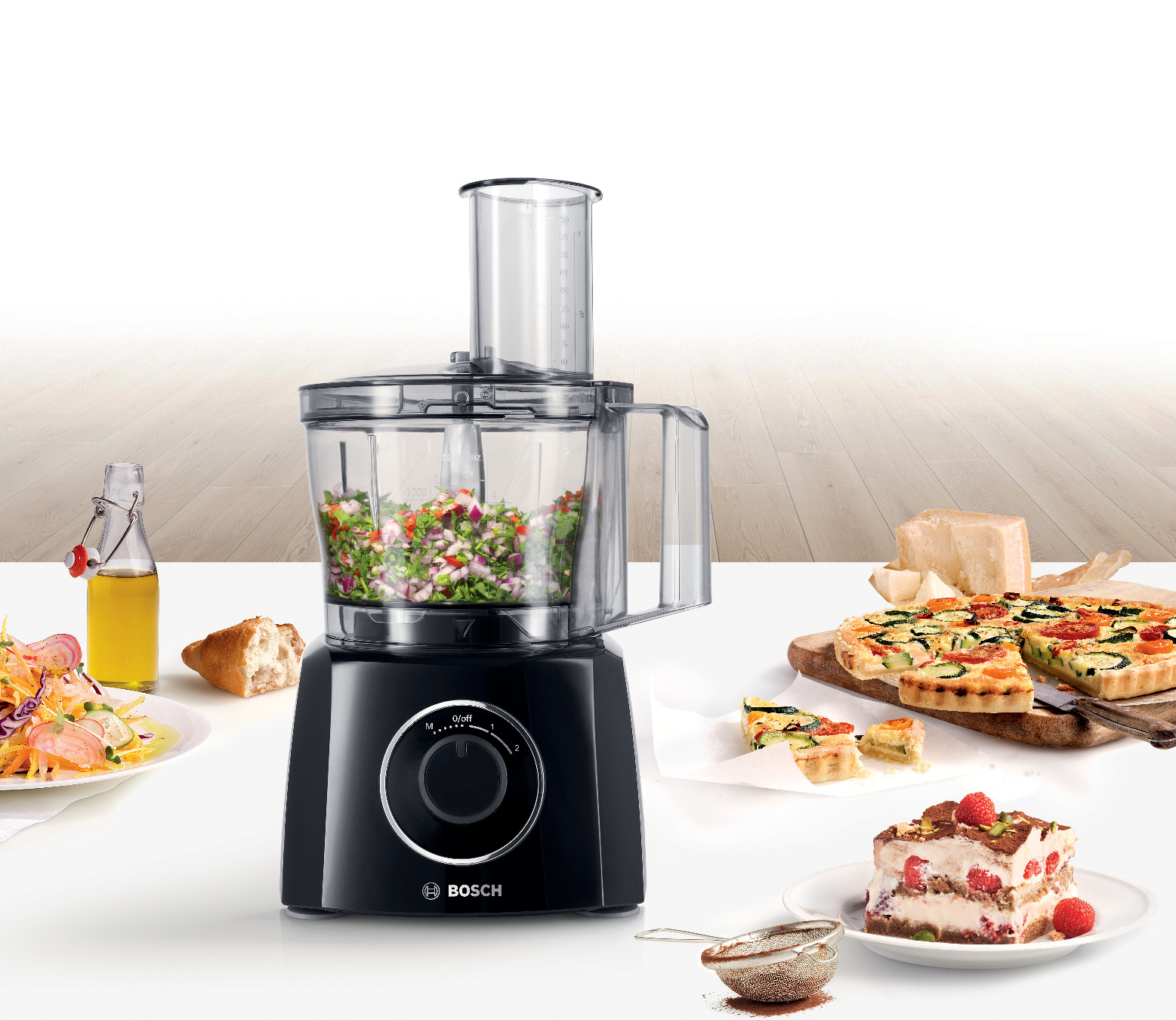 Black food processor with food and ingredients around it, on a white surface.
