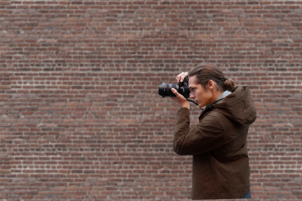 Man with camera takes photos. He is wearing a brown jacket and stands in front of a brick wall.