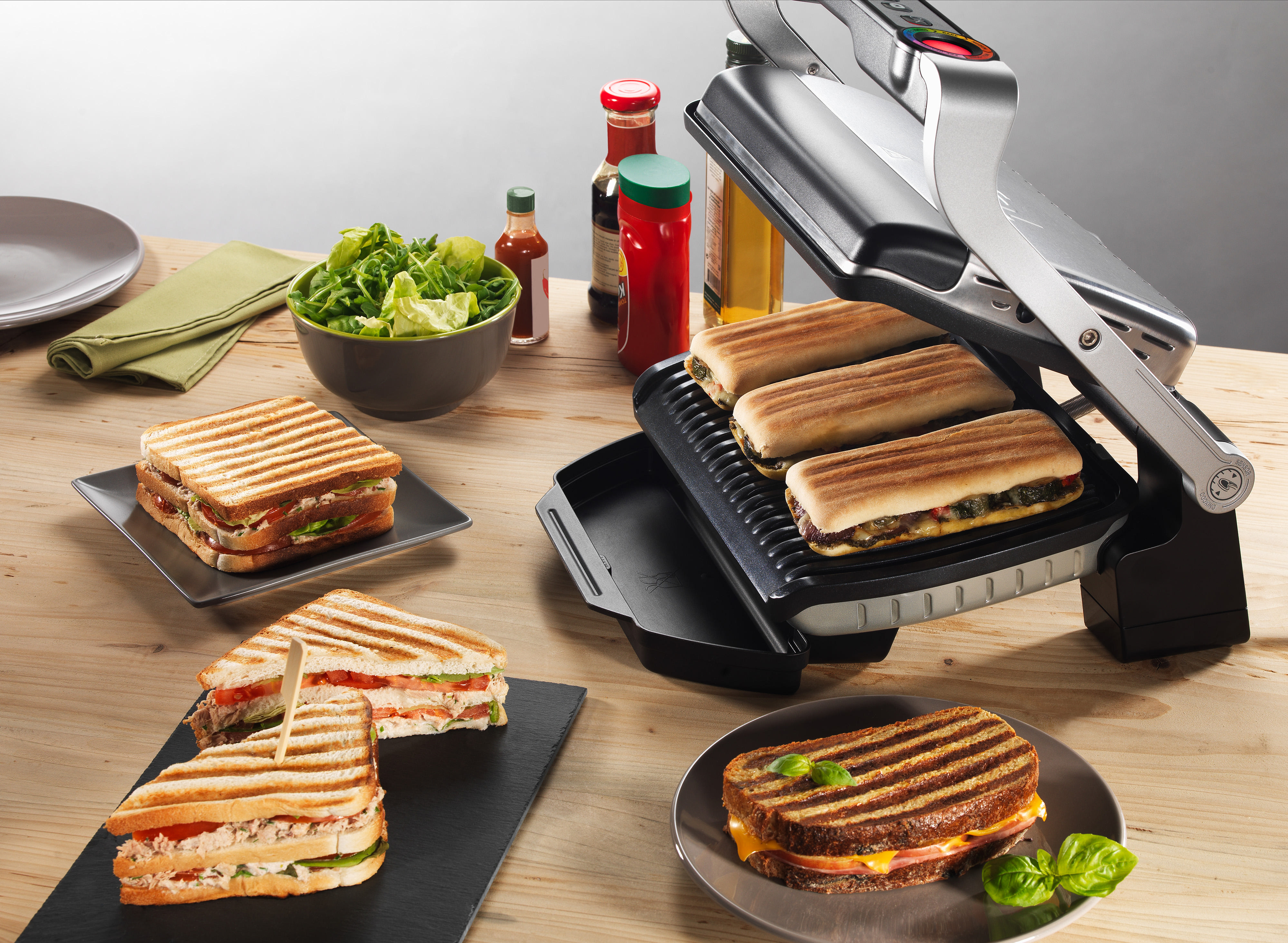 Sandwiches und eine Panini-Presse auf einem Holztisch mit Beilagen.