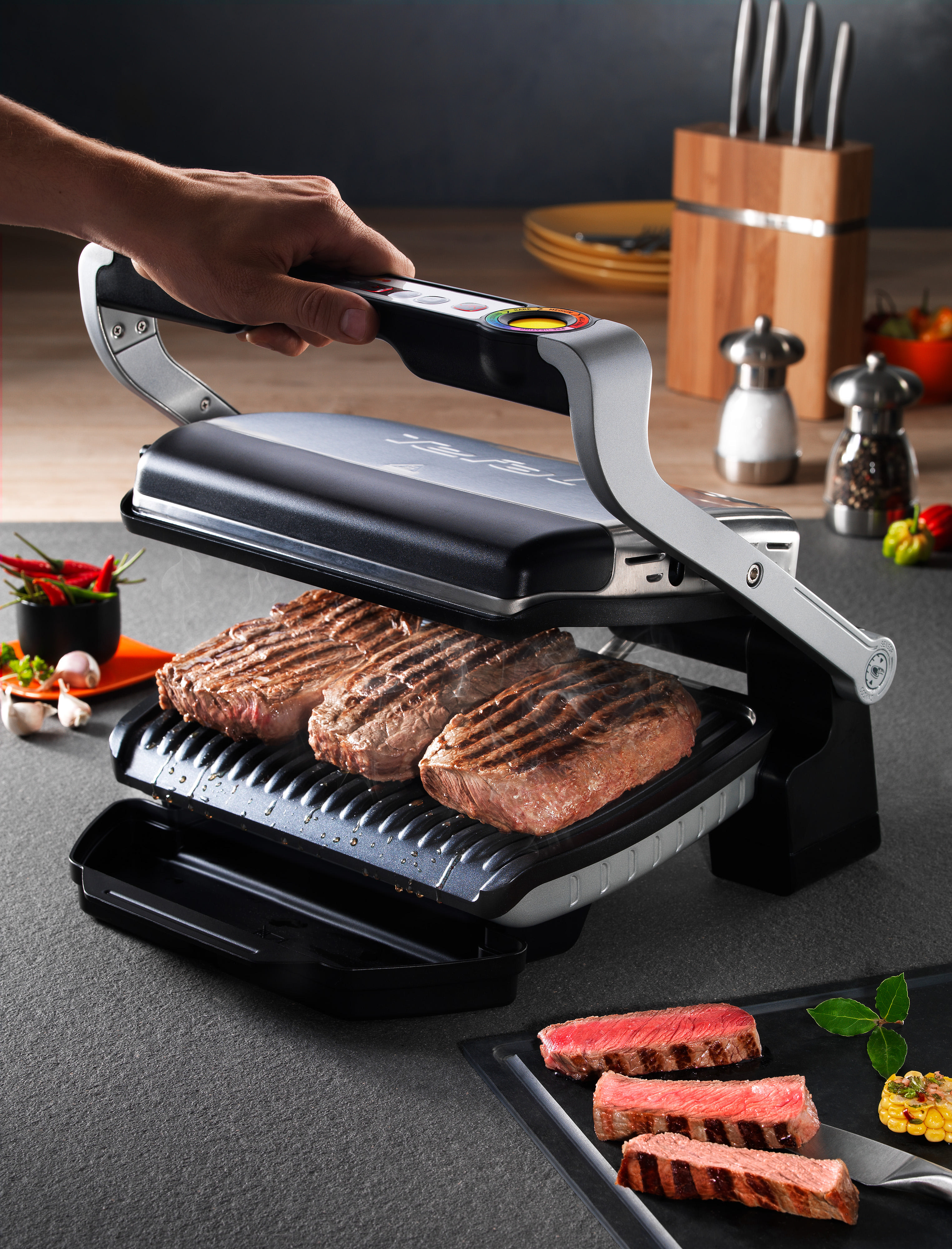 Elektrischer Grill mit bratenen Steaks. Hand schließt den Deckel. Gemüse und Gewürze sind daneben platziert.