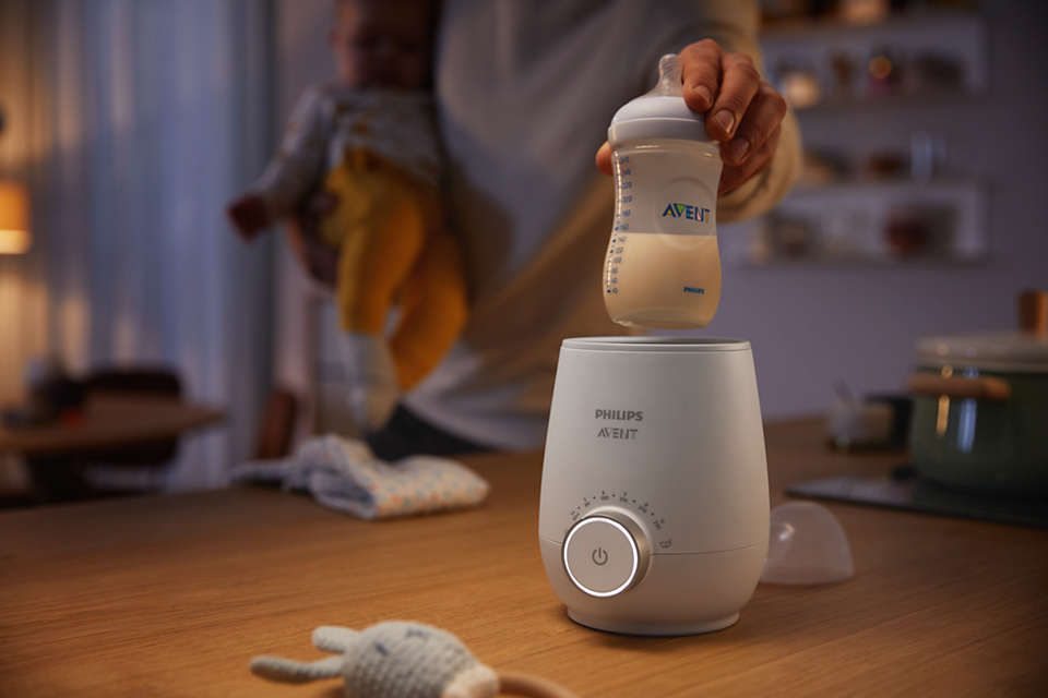 Eine Hand hält eine Avent-Milchflasche über einen weißen Philips Avent Flaschenwärmer.
