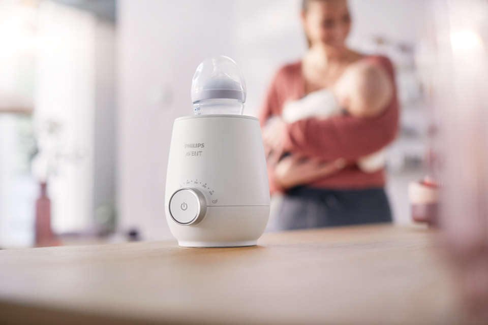Ein weißer Flaschenwärmer mit Babyflasche steht auf einem Tisch. Eine Frau hält ein Baby.