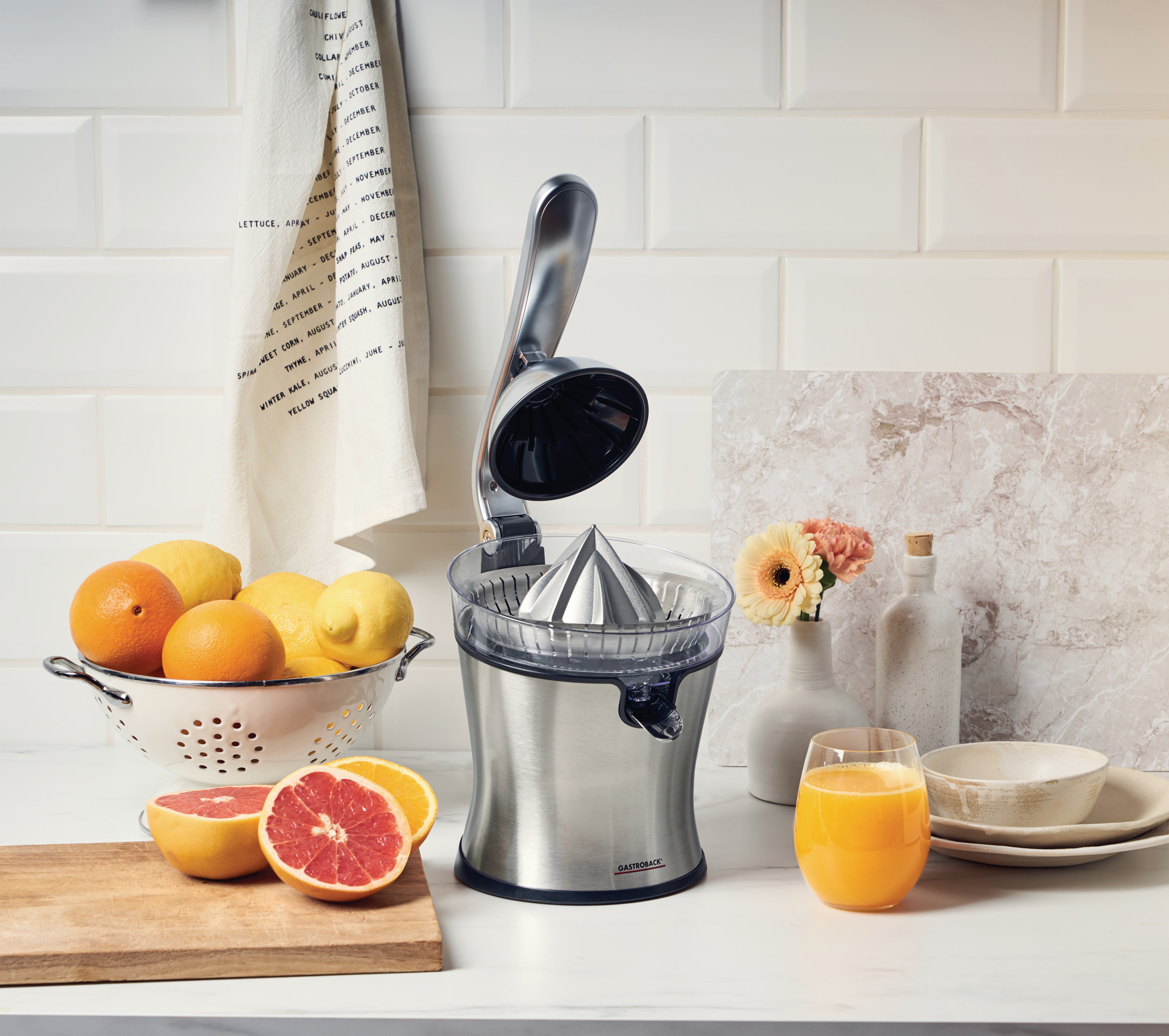 Entsafter aus Edelstahl mit Obst: Orangen, Zitronen, Grapefruit, Saft und Blumen.