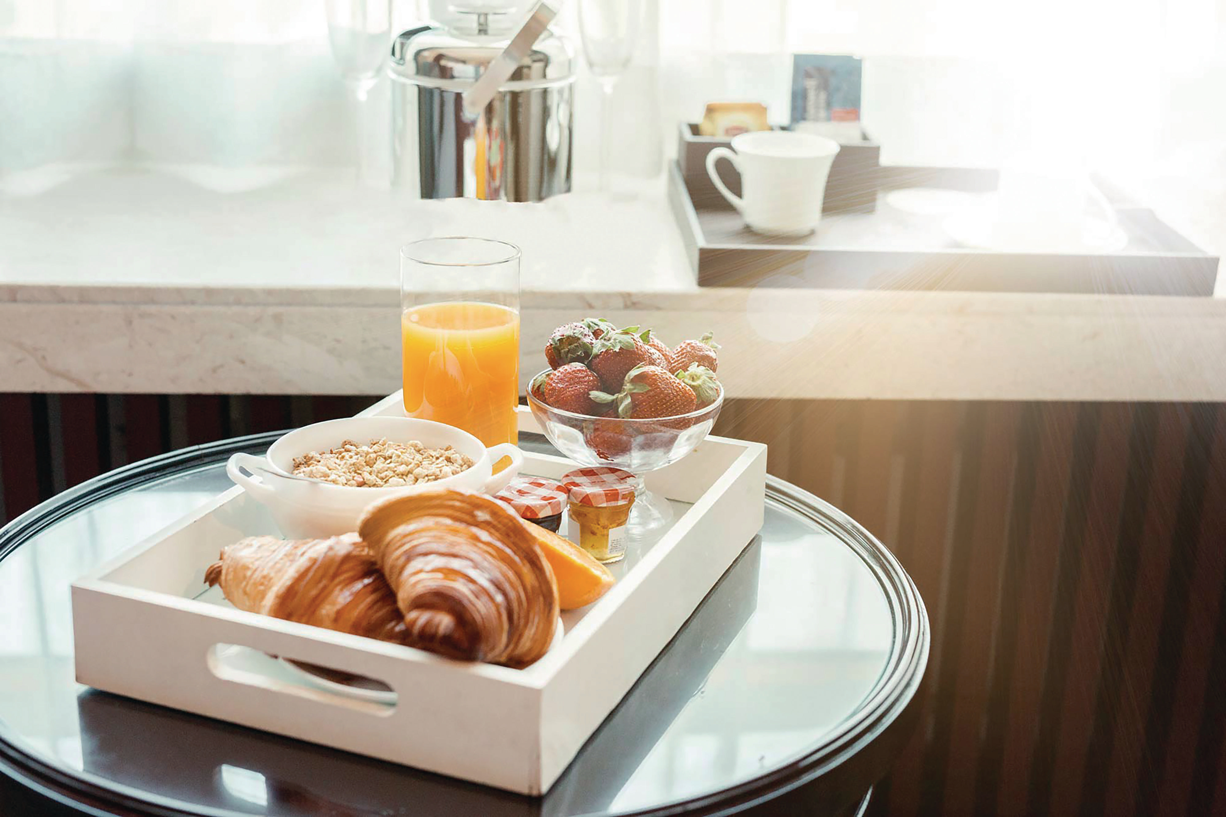 Frühstückstablett: Croissants, Obst, Saft und Müsli auf einem Glastisch.