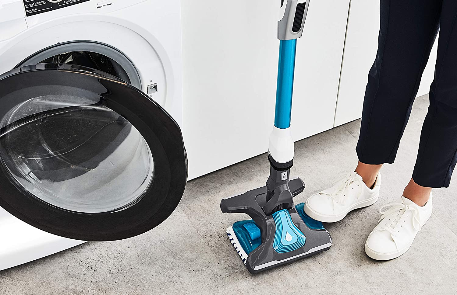 Persoon die een draadloze stofzuiger gebruikt naast een wasmachine. Witte schoenen.