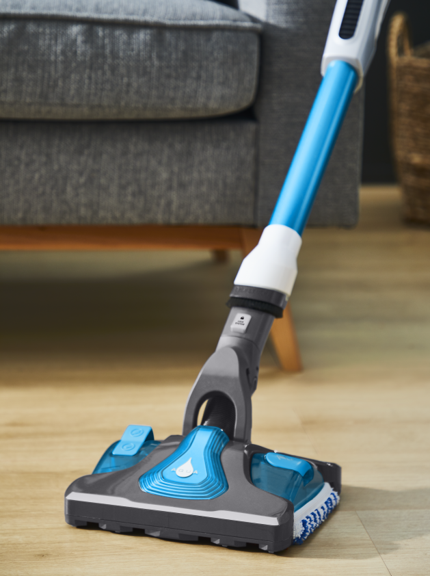 Een blauwe en grijze draadloze stofzuiger op een houten vloer. Een grijze bank staat op de achtergrond.