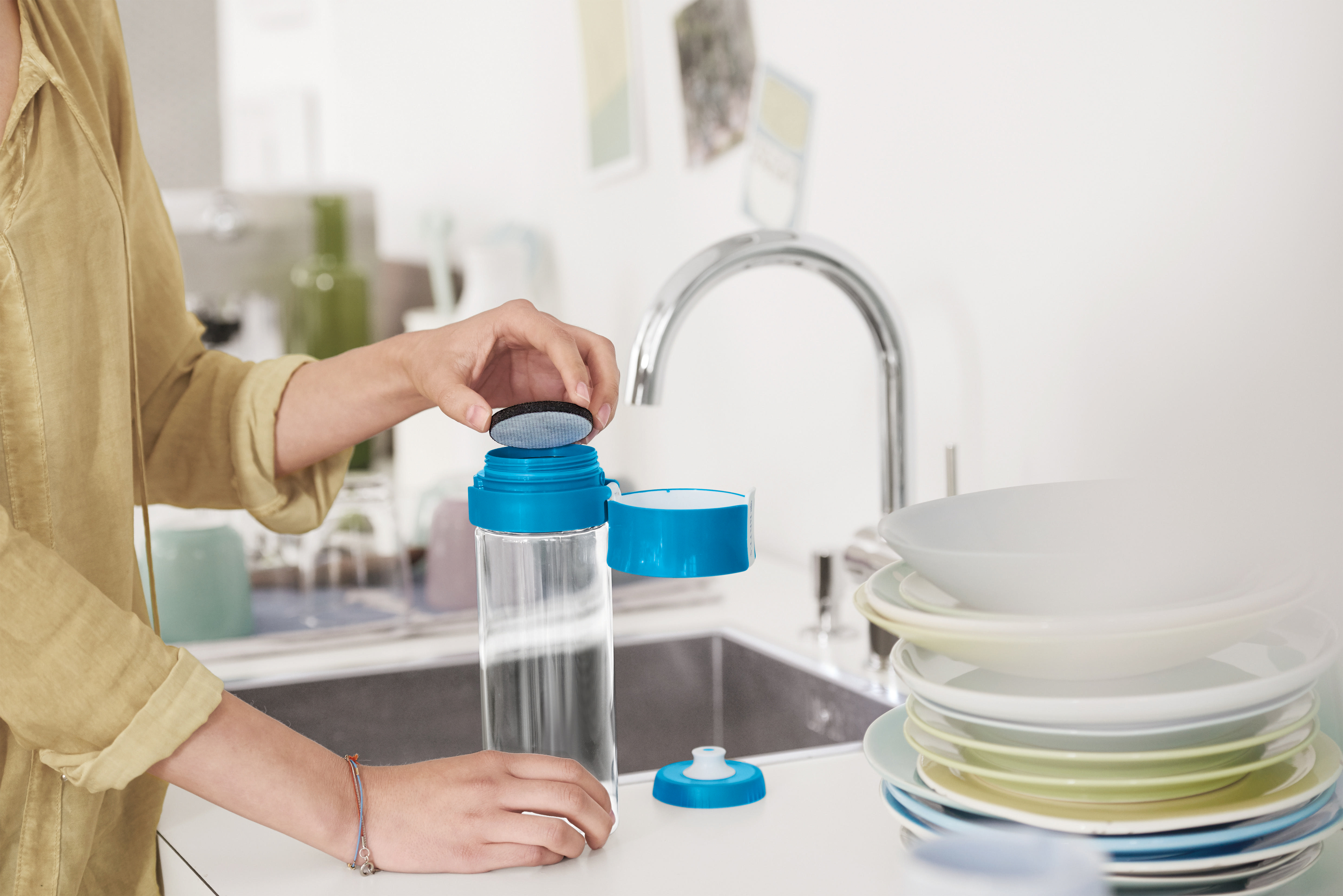 Person montiert eine Wasserflasche, mit Filter und blauem Deckel, in der Nähe von Spüle und Tellern.