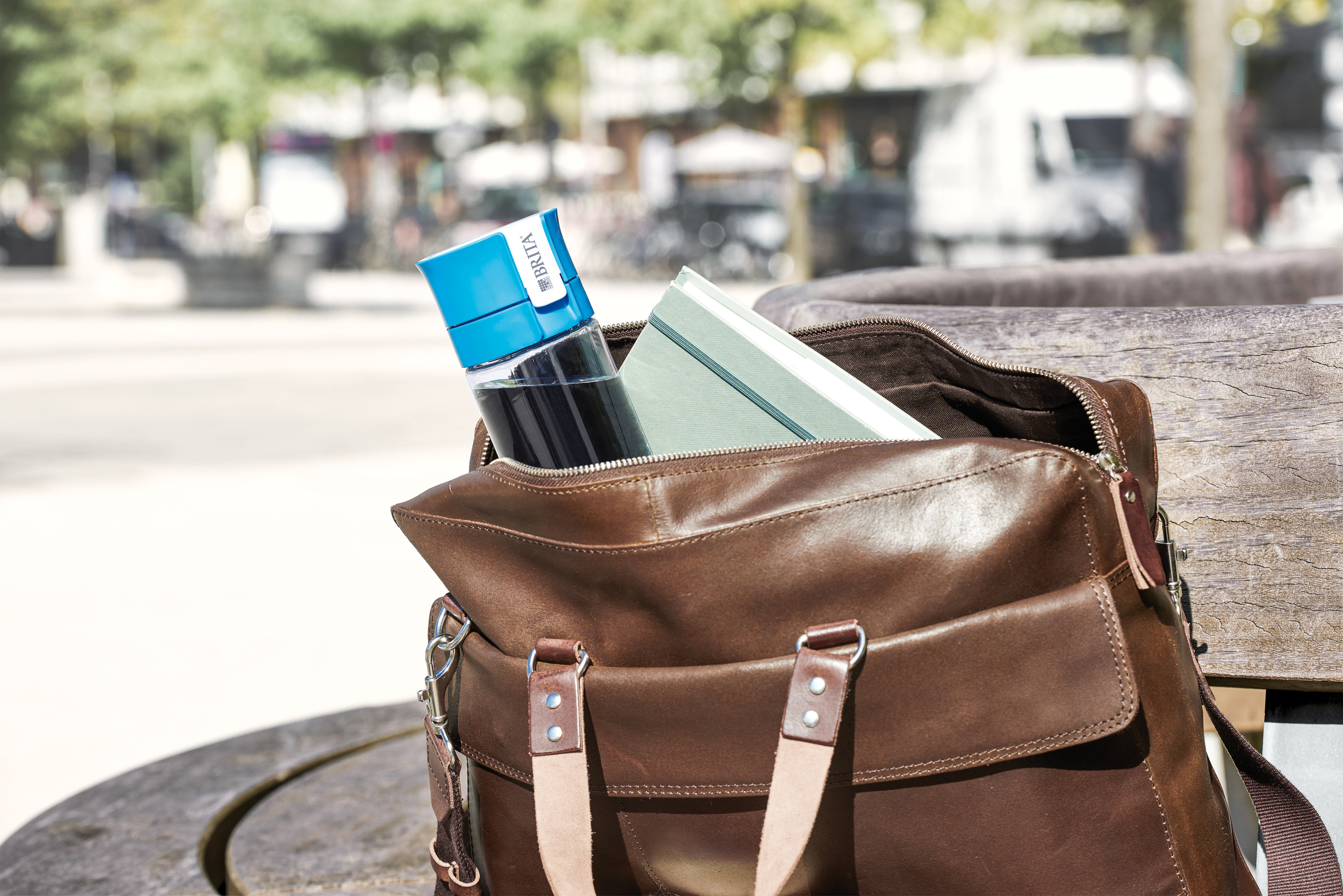 Braune Ledertasche offen auf einer Holzbank. Innen eine Wasserflasche und ein Buch. Hintergrund: Park.