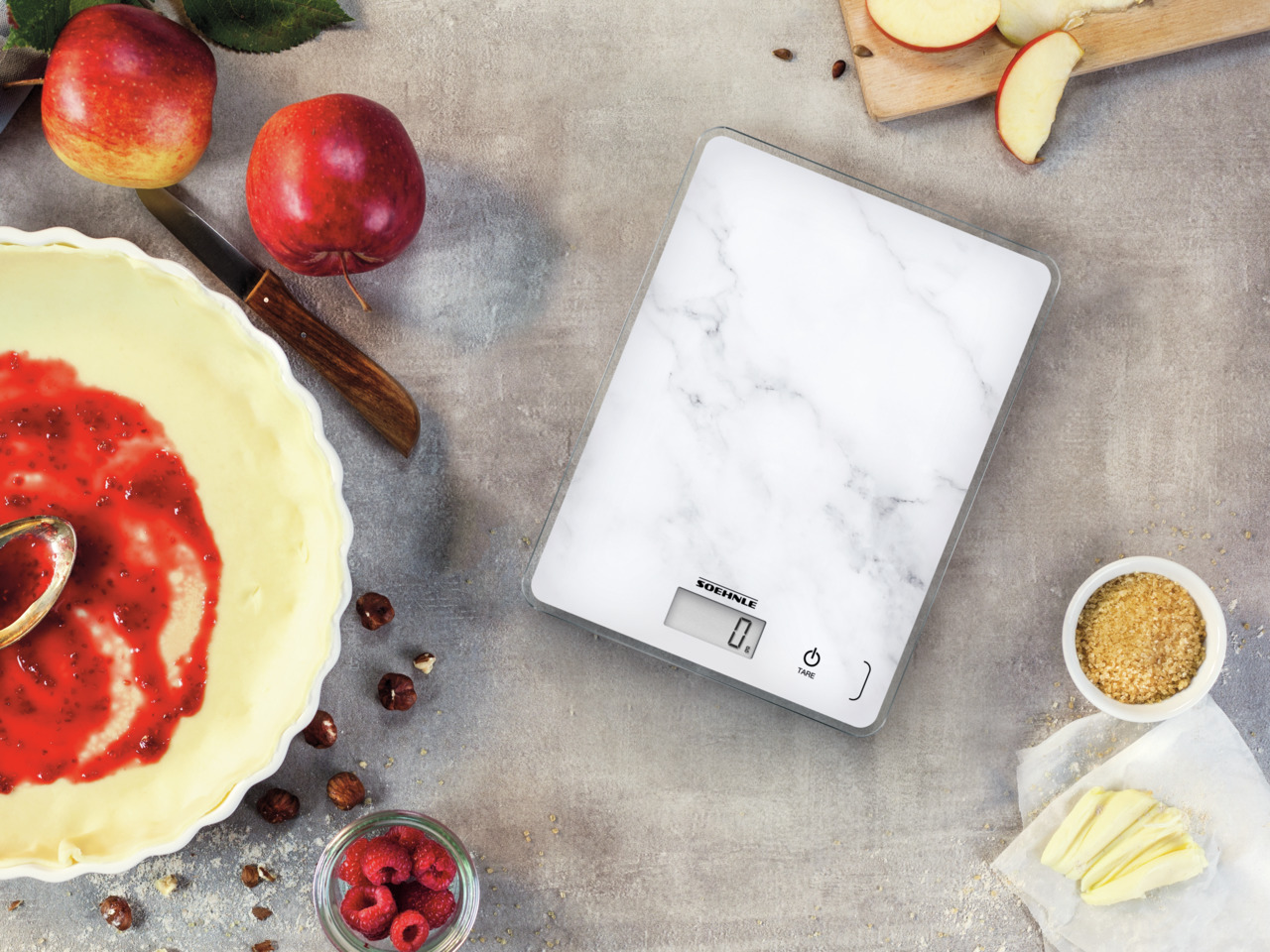 Vue aérienne. Une balance à motif marbre, des pommes, une tarte avec garniture rouge et ingrédients sur une surface grise.