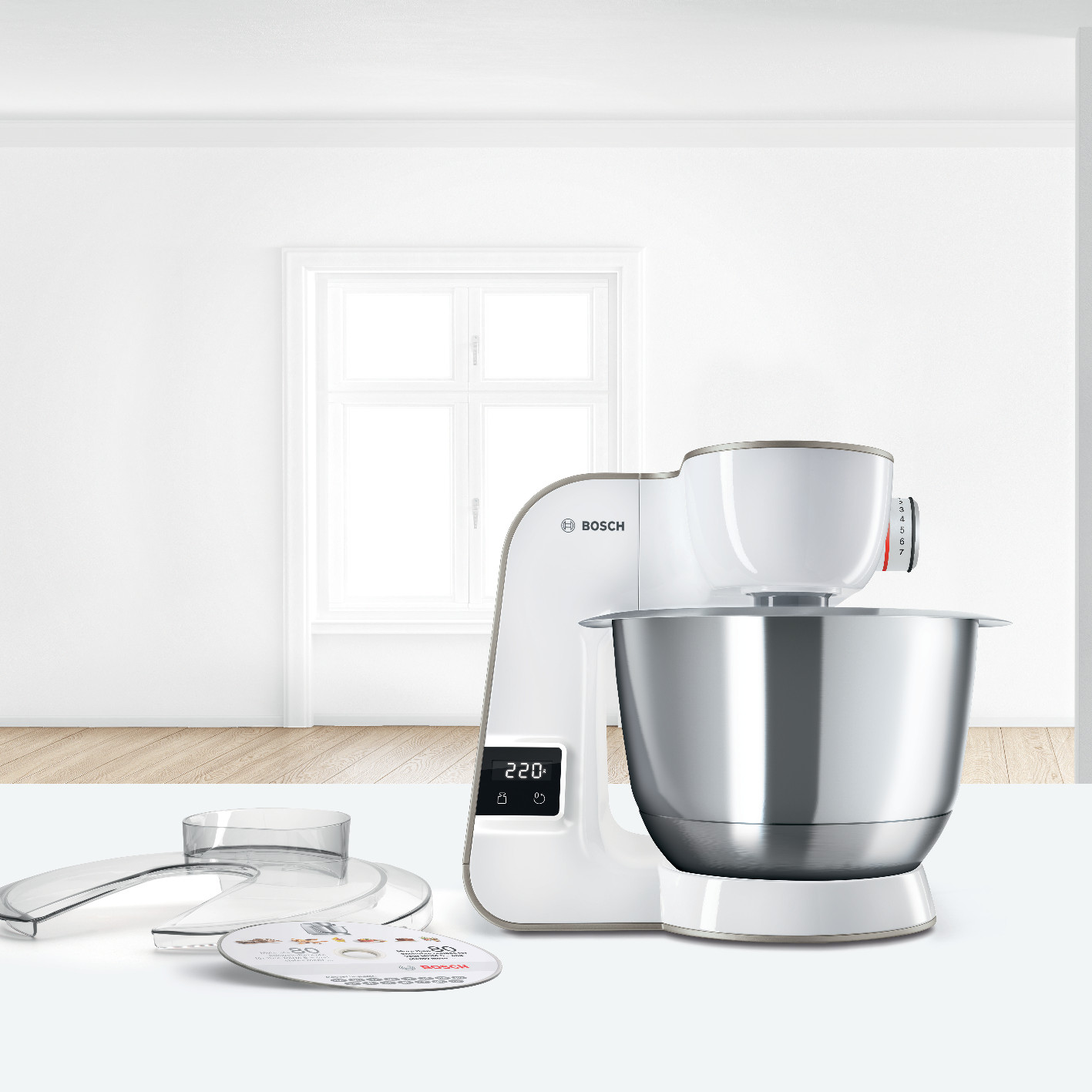Een witte keukenmachine met roestvrijstalen kom en accessoires op een witte tafel.