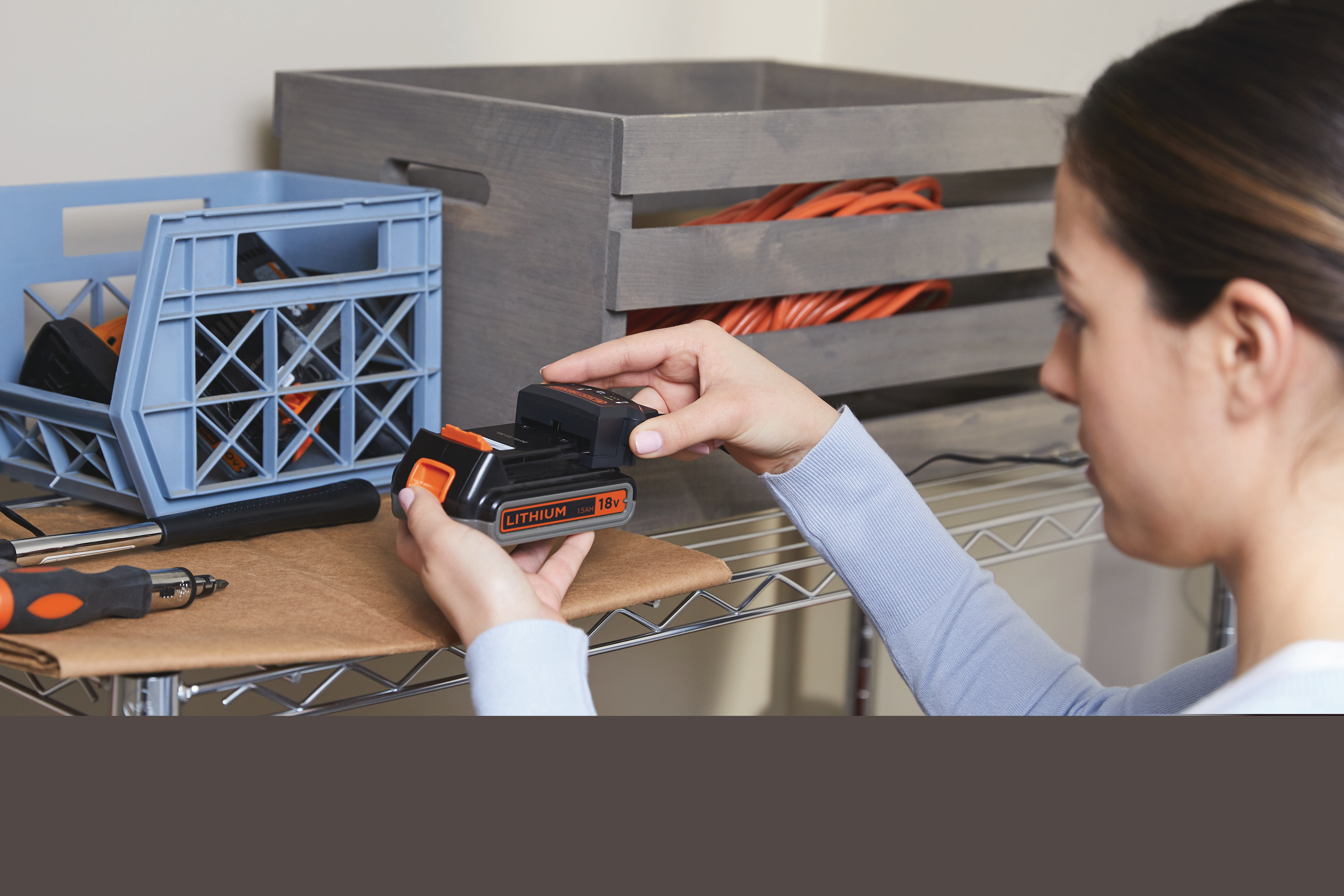 Frau, die einen schwarzen Akku in ein schwarz-oranges Black+Decker-Werkzeug einsetzt. Regale und Kisten im Hintergrund.