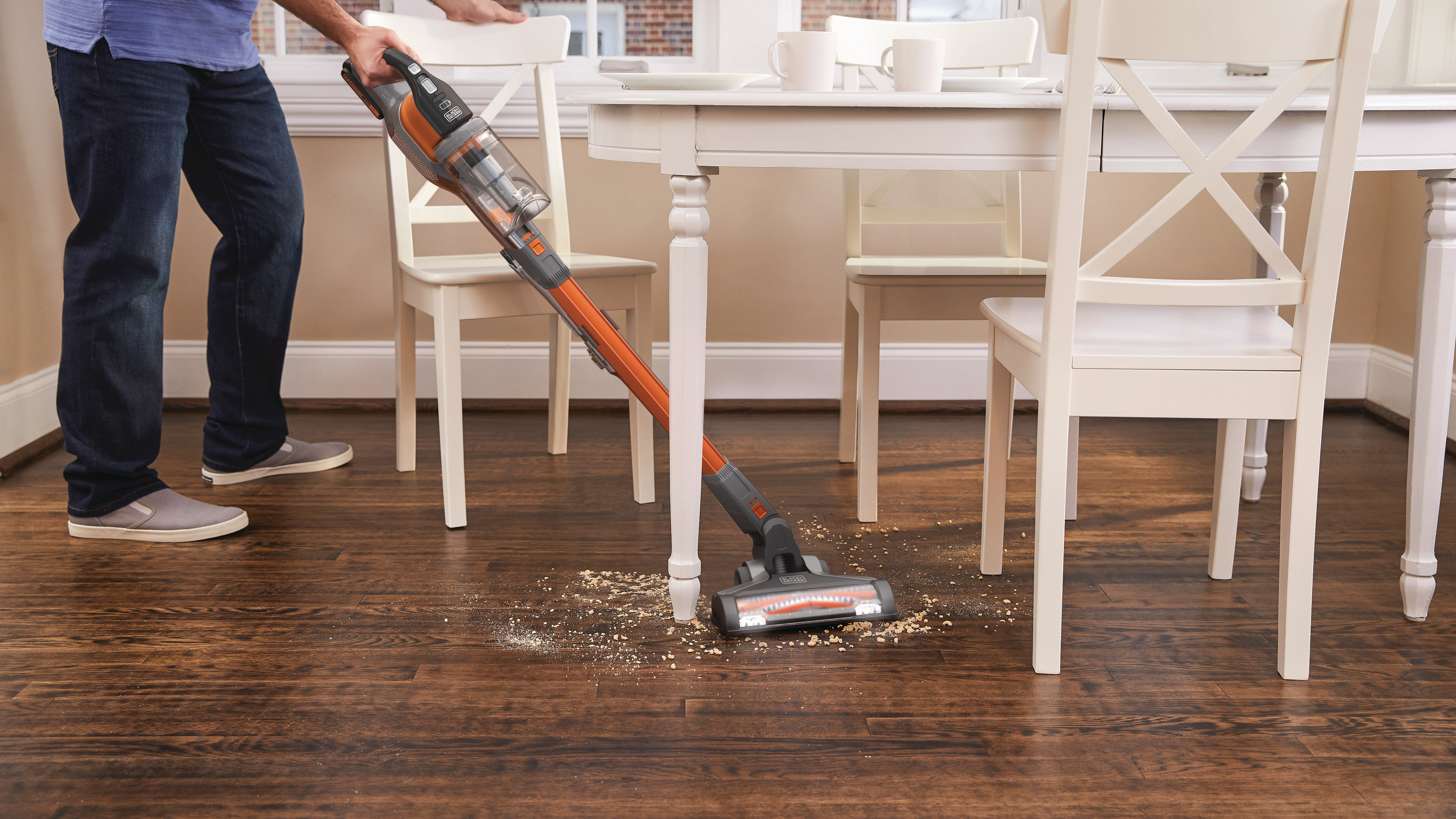 Eine Person saugt einen Holzboden. Ein kabelloser Staubsauger wird verwendet, und es gibt einen Holztisch.