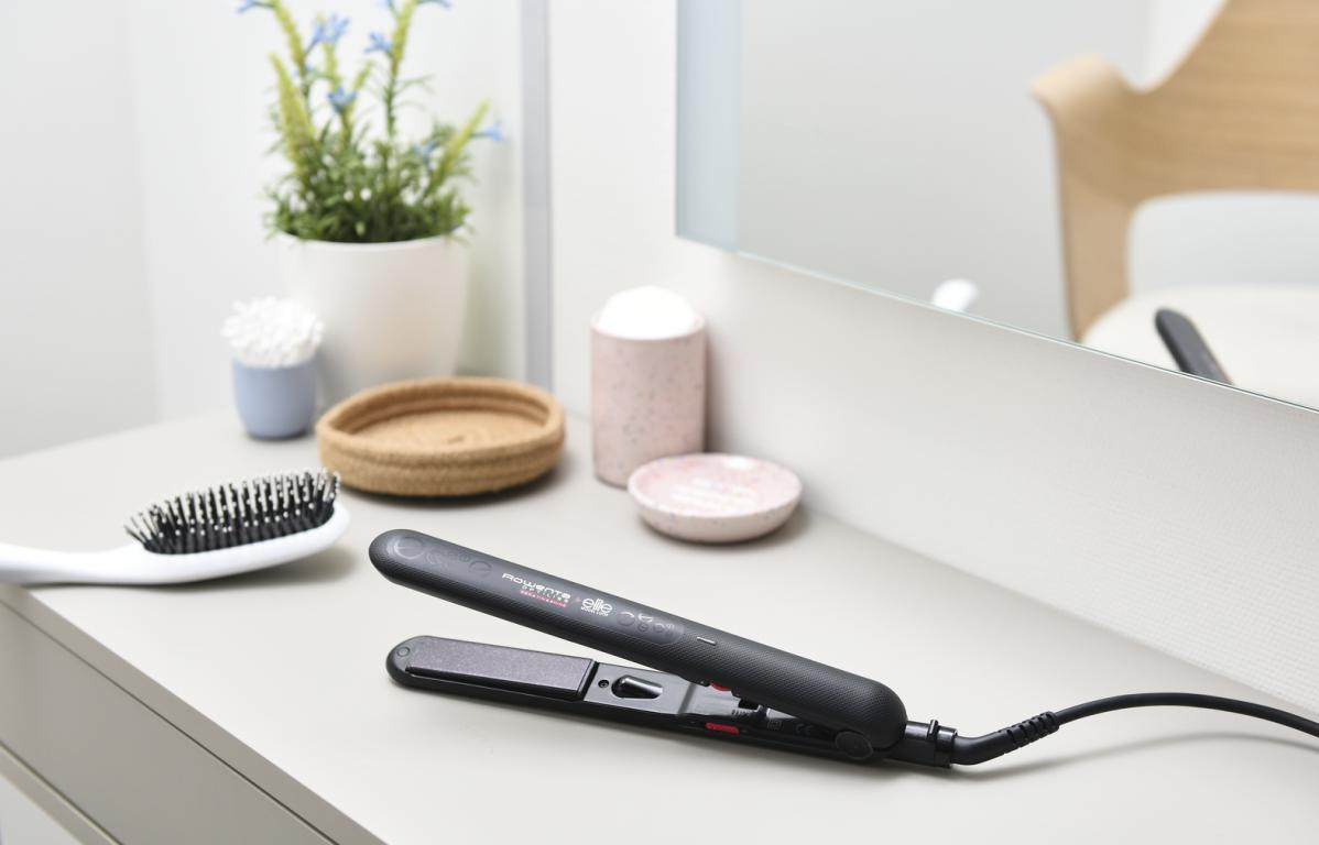 Una plancha para el pelo sobre una mesa gris, con un cepillo y cosméticos.