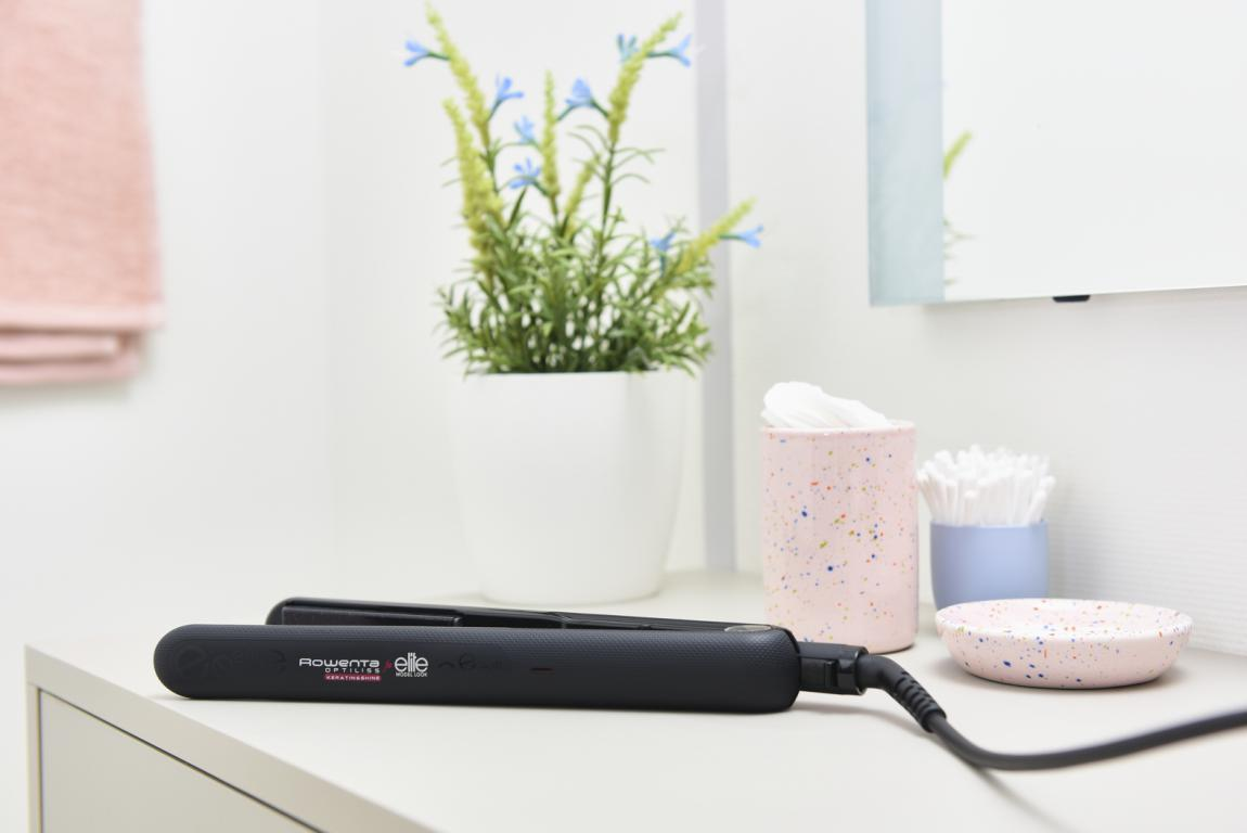 Una plancha de pelo negra sobre una superficie blanca, con accesorios de baño en el fondo.