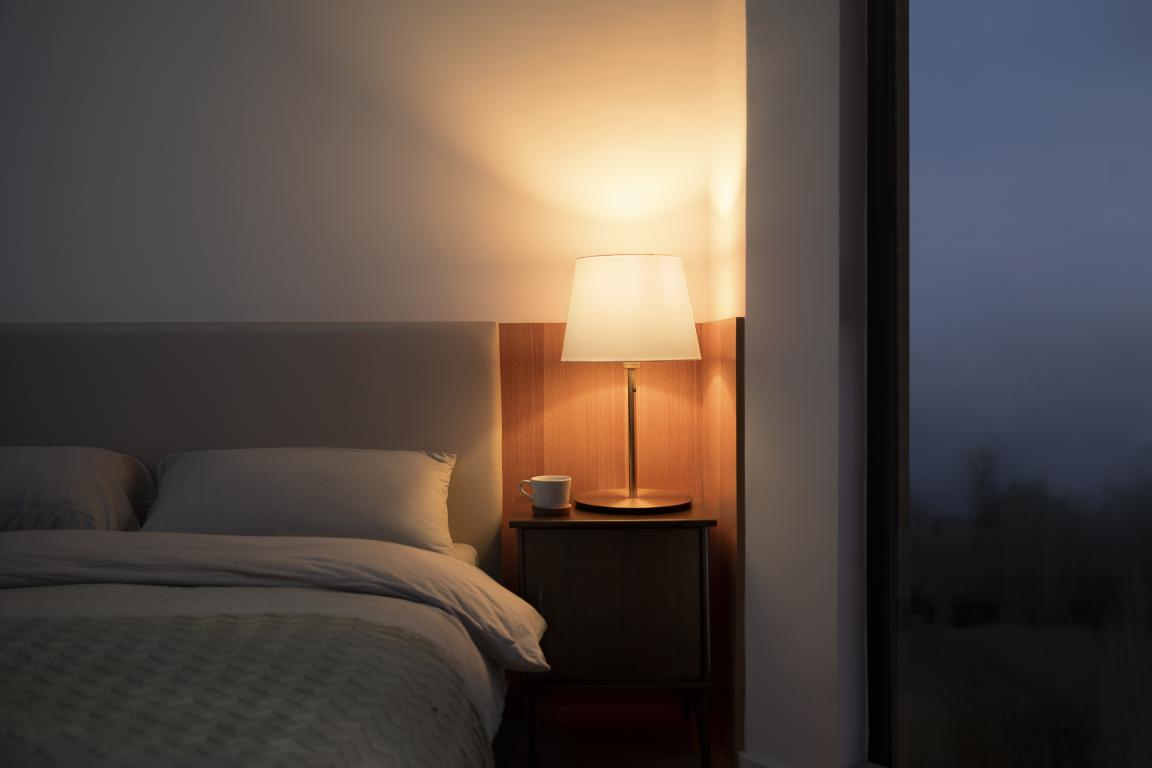 Una escena de dormitorio con una lámpara en una mesita de noche y una cama. Es de poca luz.