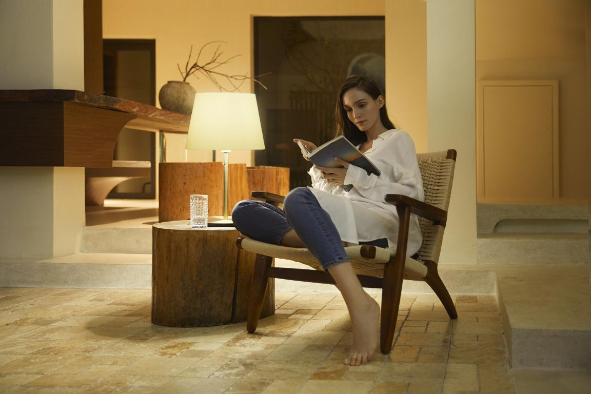 Mujer sentada en una silla, leyendo un libro. Mesa con vaso y lámpara cerca.