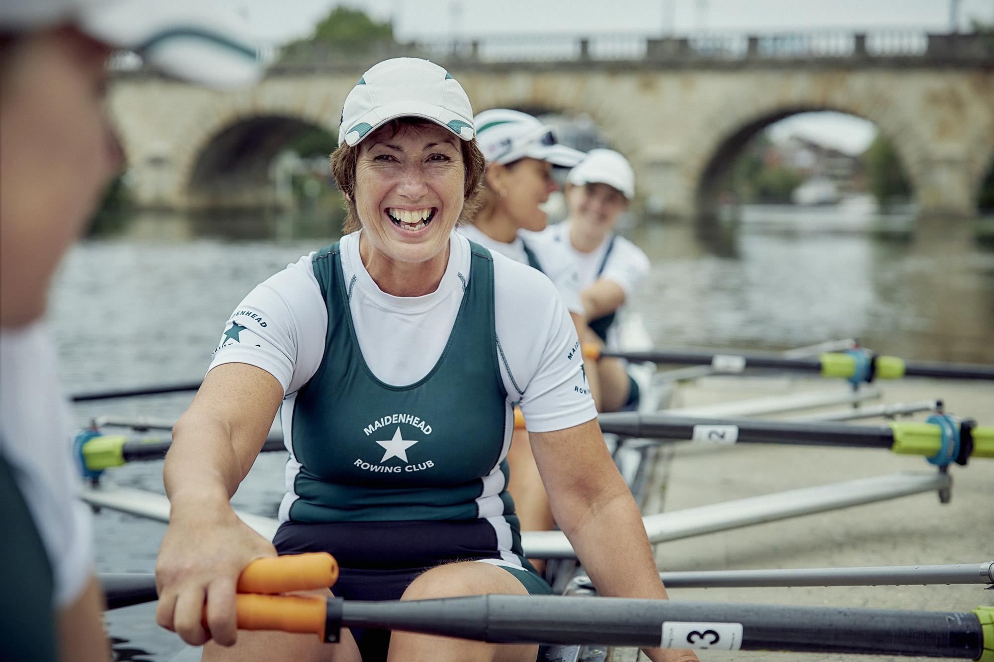 Eine Frau in einem Ruderboot, breit lächelnd, mit Teamkollegen und Brücke im Hintergrund.