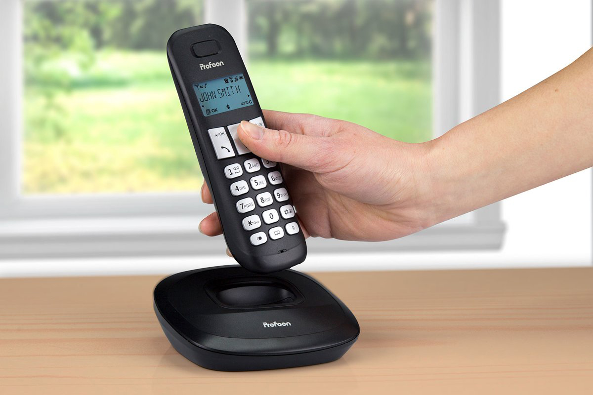 Un téléphone sans fil noir sur une base de chargement, tenu par une main, devant une fenêtre.
