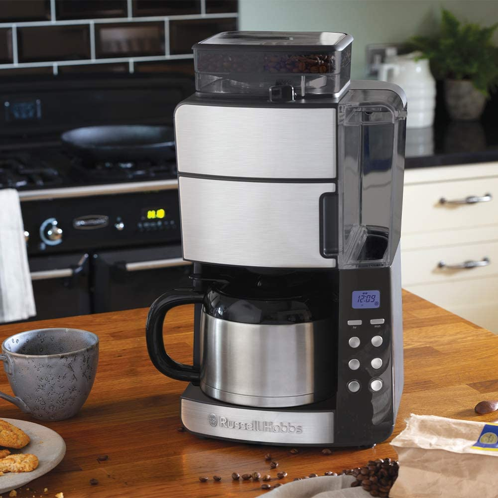 Egy rozsdamentes acél Russell Hobbs kávéfőző egy fa pulton. Balra csésze és keksz. Konyhai háttér.
