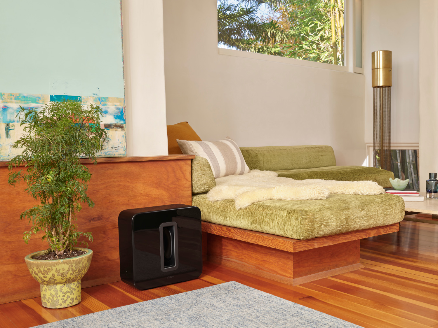 Een woonkamer met een bank, een plant en een subwoofer. Het heeft houten vloeren.
