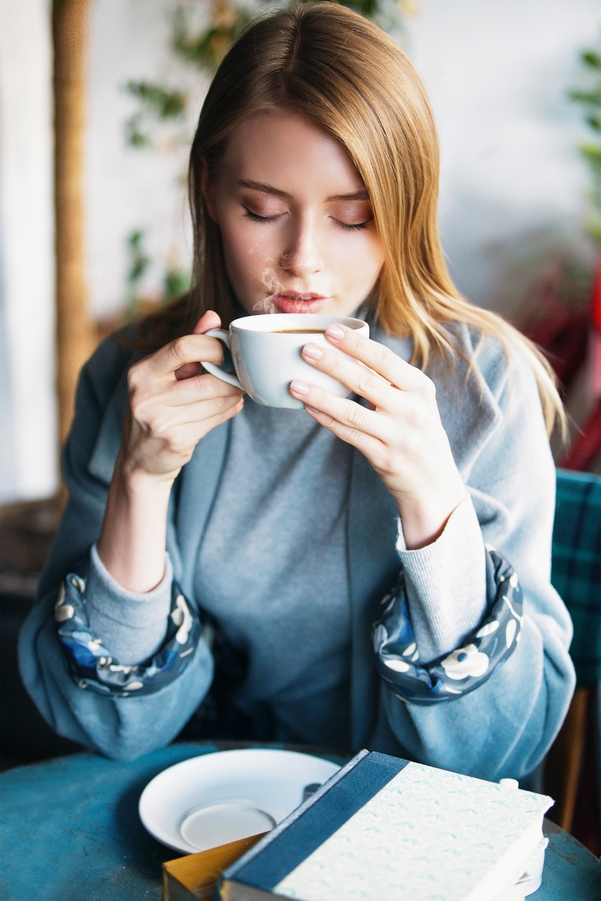 Eine Frau mit blonden Haaren trinkt Kaffee in einem Café mit einem Buch auf dem Tisch.