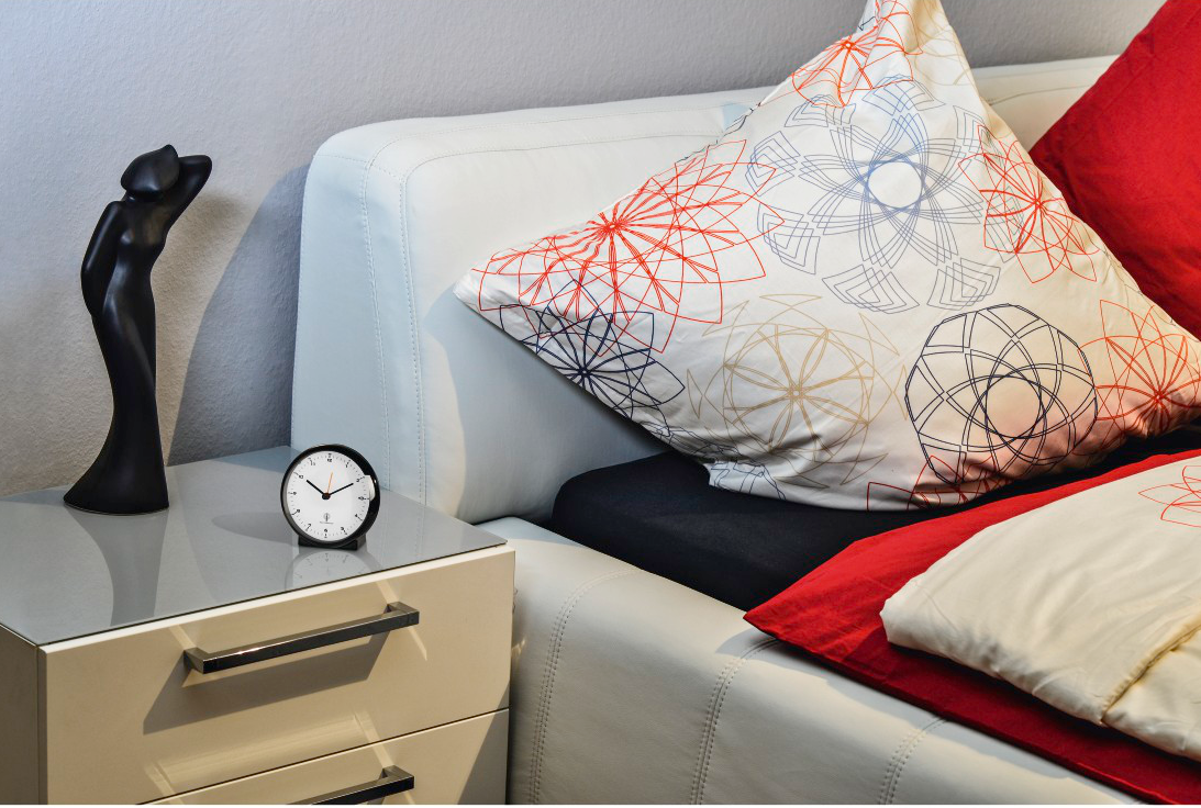 Nachttisch mit Uhr und Statue. Kissen auf dem Bett mit roten und gemusterten Bezügen.