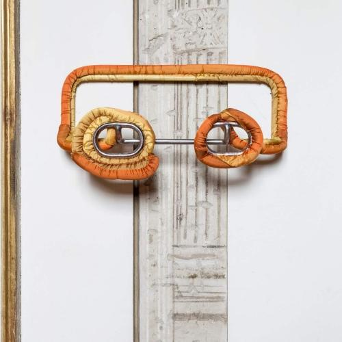 Œuvre d'art en tissu orange sur fond blanc. Elle a une forme rectangulaire incurvée avec des accents métalliques.