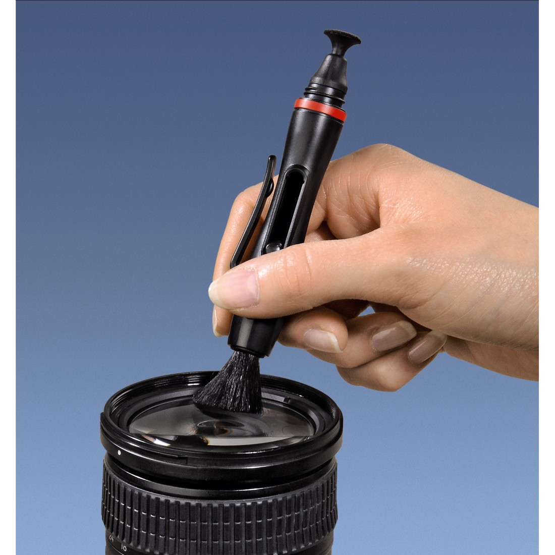 Hand hält einen schwarzen Objektiv-Reinigungsstift über einer Kamera-Linse vor blauem Hintergrund.