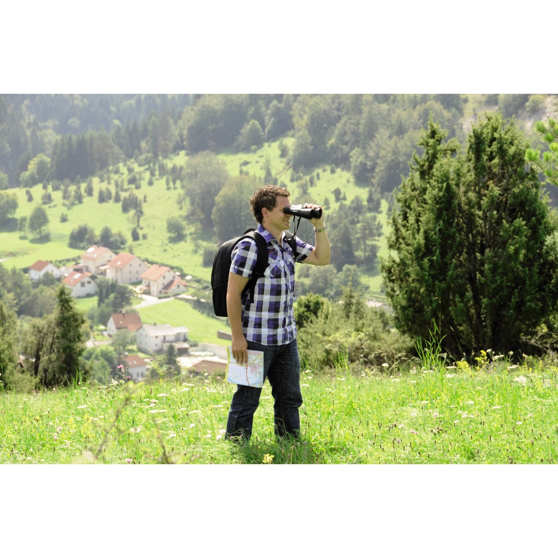 Ein Mann im karierten Hemd blickt mit einem Fernglas auf einer grünen Wiese mit einer Karte.
