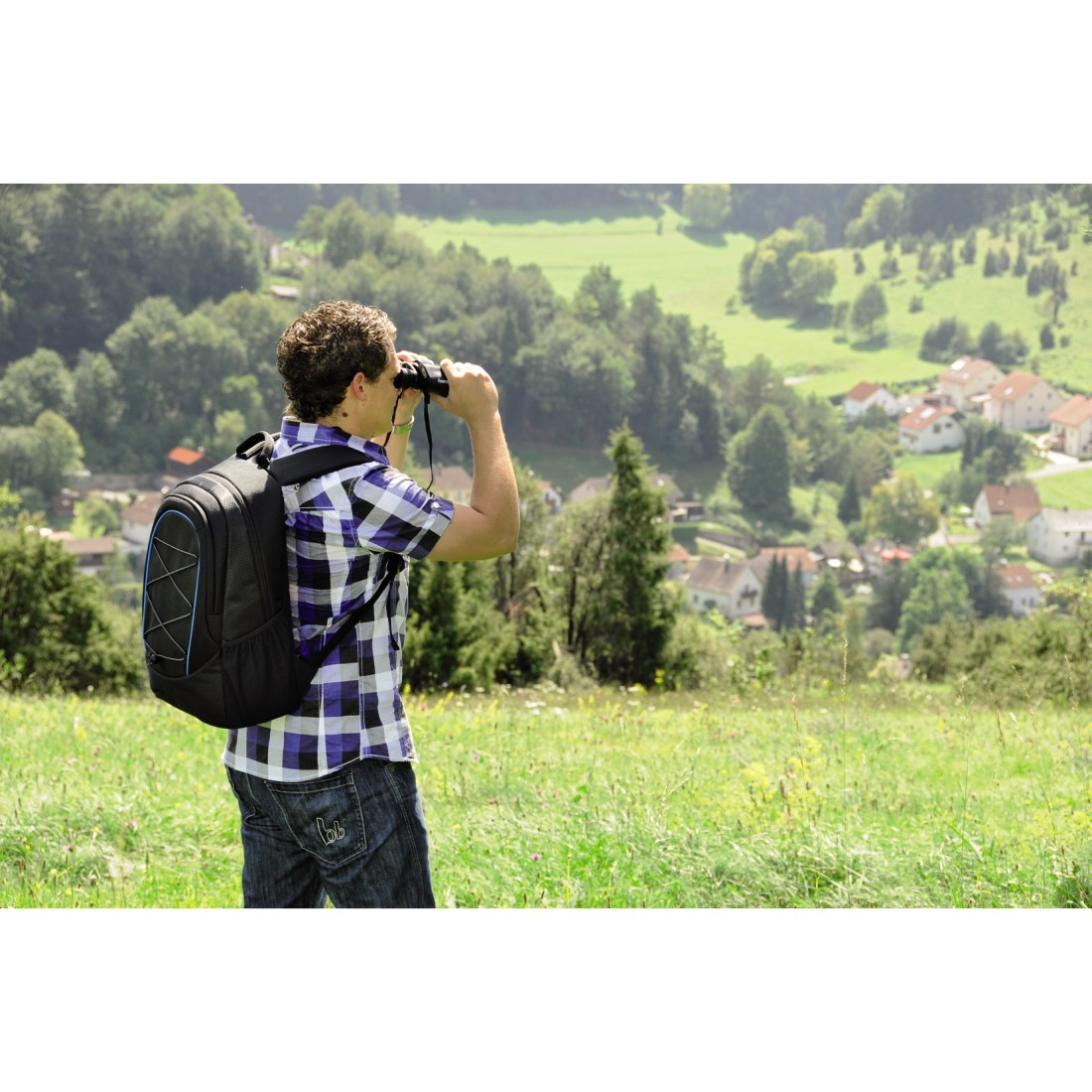 Mann mit Rucksack, der durch ein Fernglas auf einem grasbewachsenen Hügel blickt. Bäume und Häuser im Hintergrund.